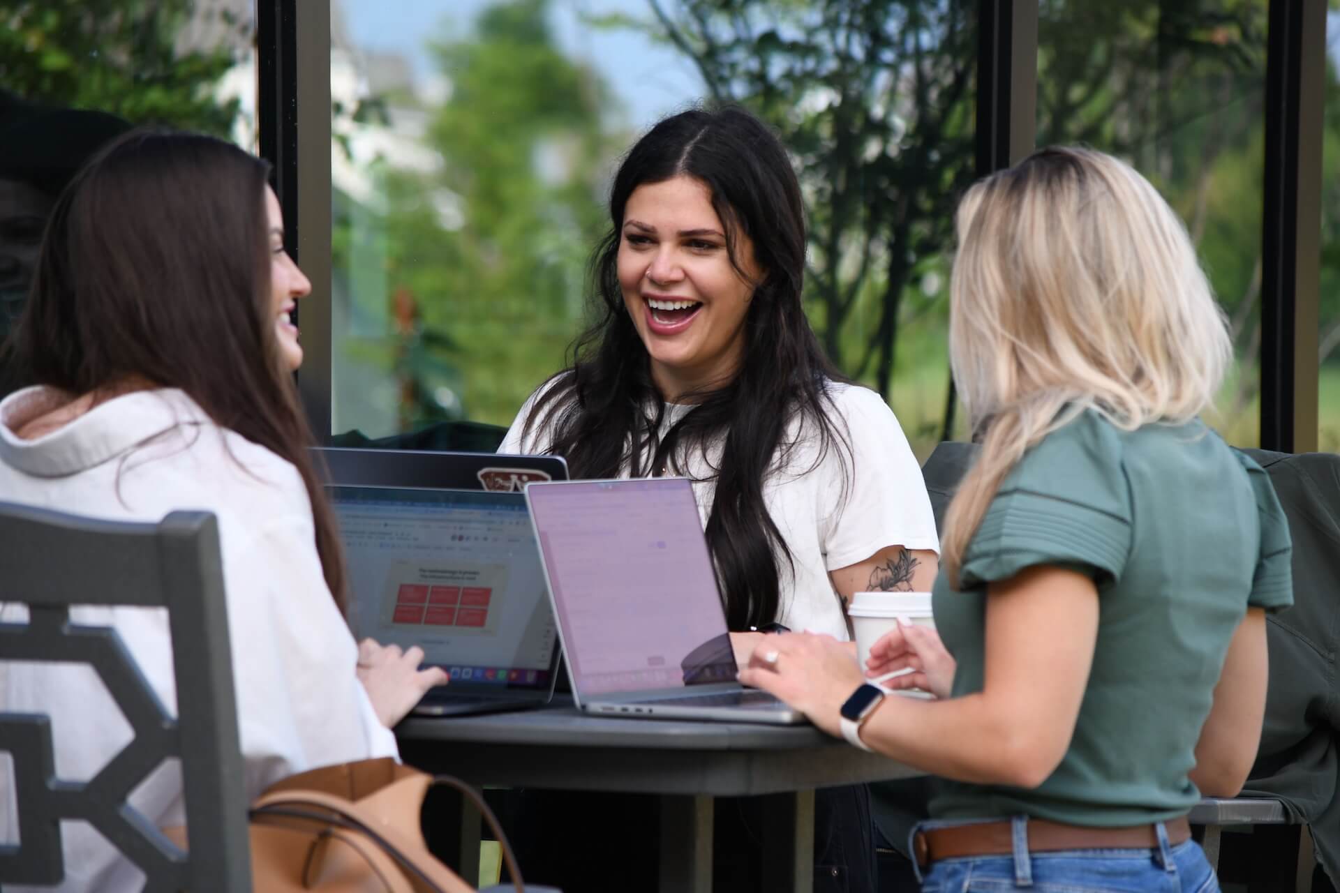 DSC_8185 Three women sit at an outdoor table, each with a laptop, smiling and talking. Lush greenery is visible in the background, suggesting a relaxed and pleasant atmosphere.