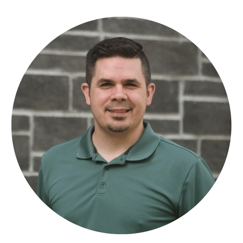 evan-cox-2026 (1) A man with short dark hair and a trimmed beard wears a green polo shirt and smiles in front of a gray brick wall.