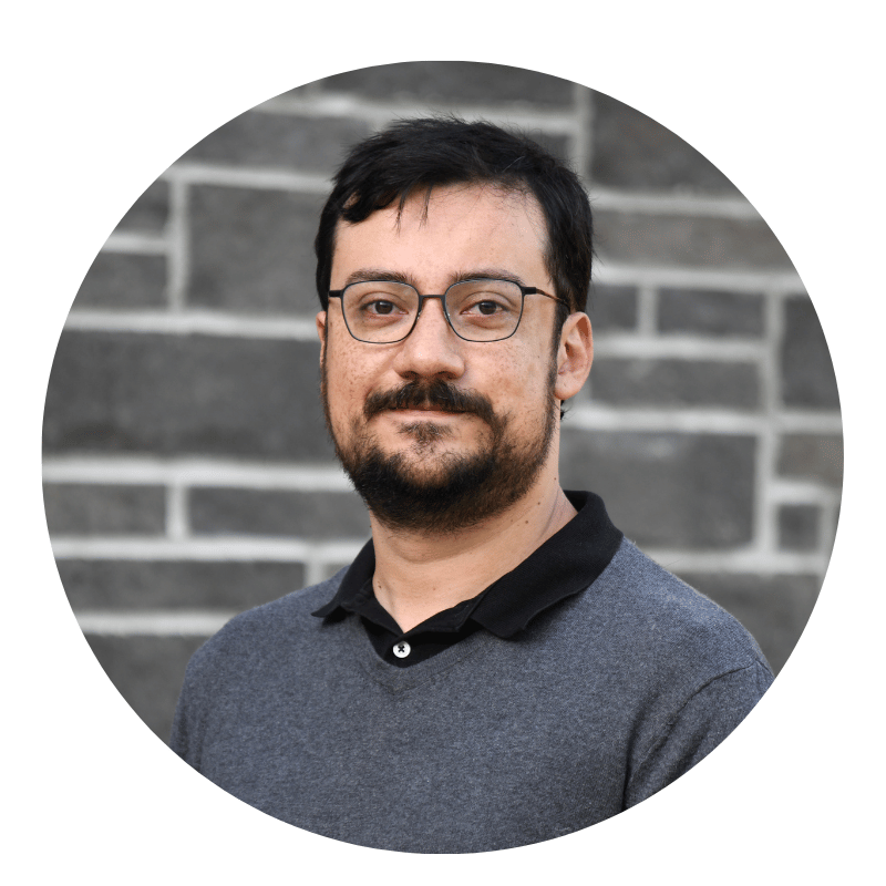 germán-garcía-2026 A person with short dark hair, beard, and glasses, wearing a gray collared shirt, stands in front of a gray brick wall, looking at the camera and smiling slightly.