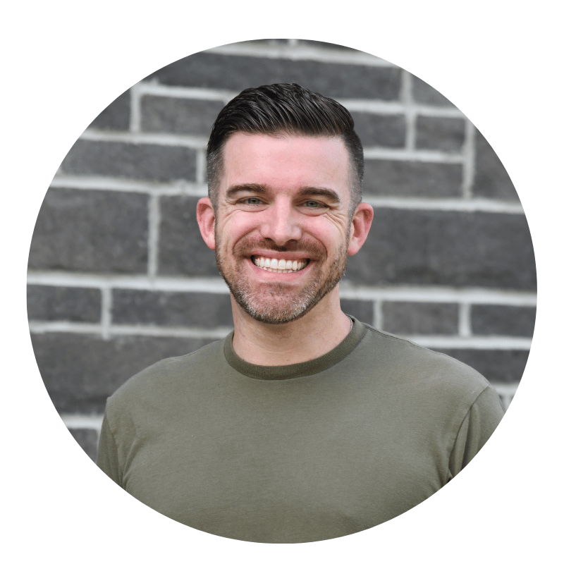 jim-gilkey-2026 A smiling man with short dark hair and a trimmed beard, wearing a green t-shirt, stands in front of a gray brick wall.