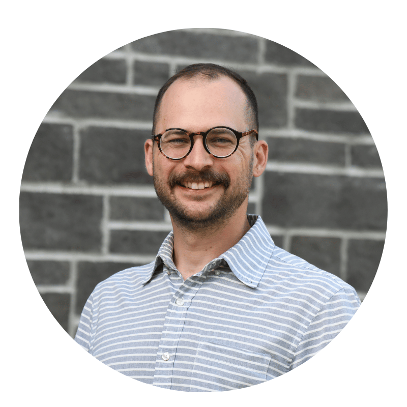 joseph-lewin-2026 A person with glasses, a mustache, and a beard, wearing a light gray striped shirt, is smiling in front of a gray brick wall.
