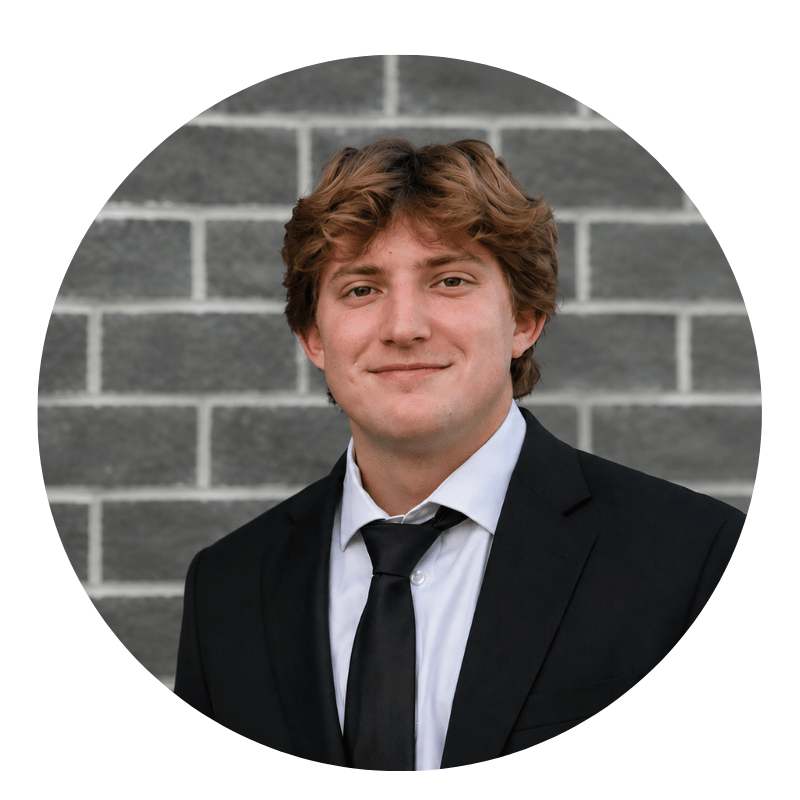 kyle-howard-2026 A young man in a black suit and tie smiles in front of a gray brick wall.