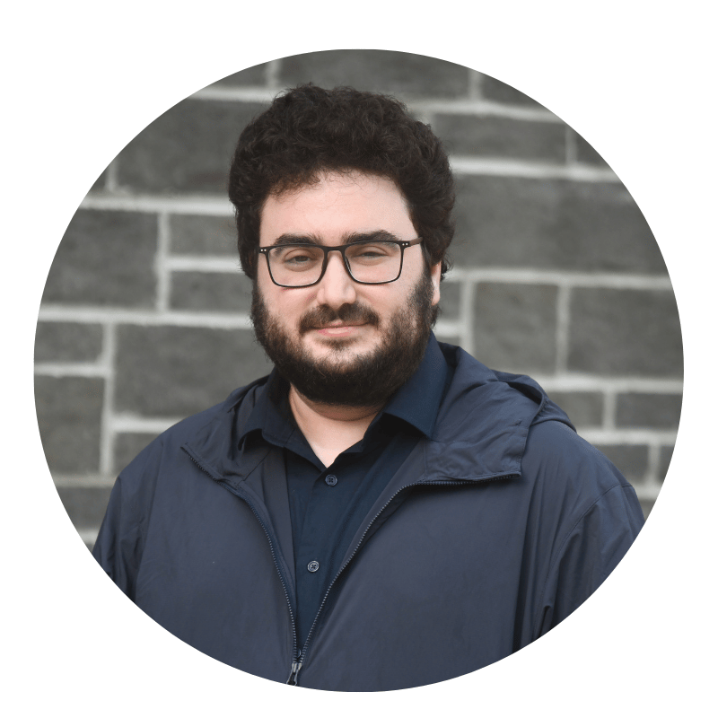mati-rosenberg-2026 A person with curly dark hair, glasses, and a beard is wearing a navy jacket and dark shirt, standing in front of a gray brick wall.
