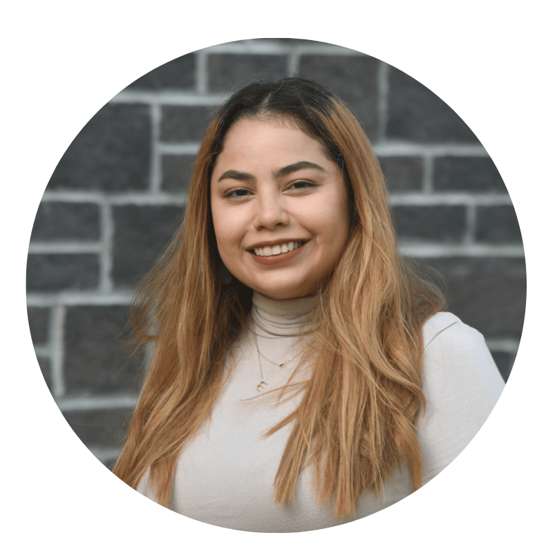 priscilla-caldera-2026 A woman with long, light brown hair smiles while standing in front of a gray brick wall, wearing a beige turtleneck top and layered necklaces.