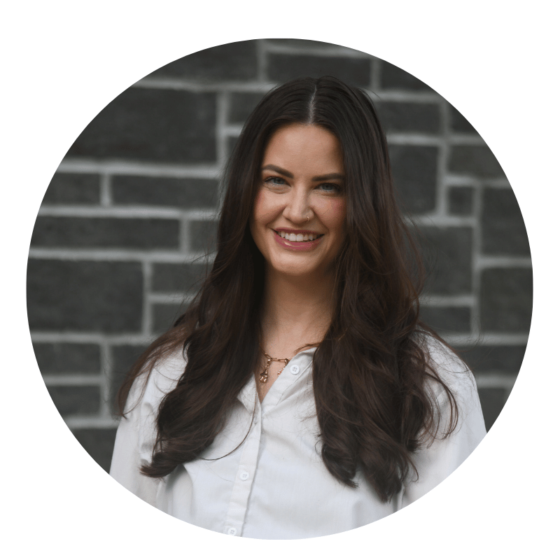 tori-stubbs-2026 Woman with long brown hair and a white shirt smiles in front of a dark gray brick wall. The photo is framed in a circular border.
