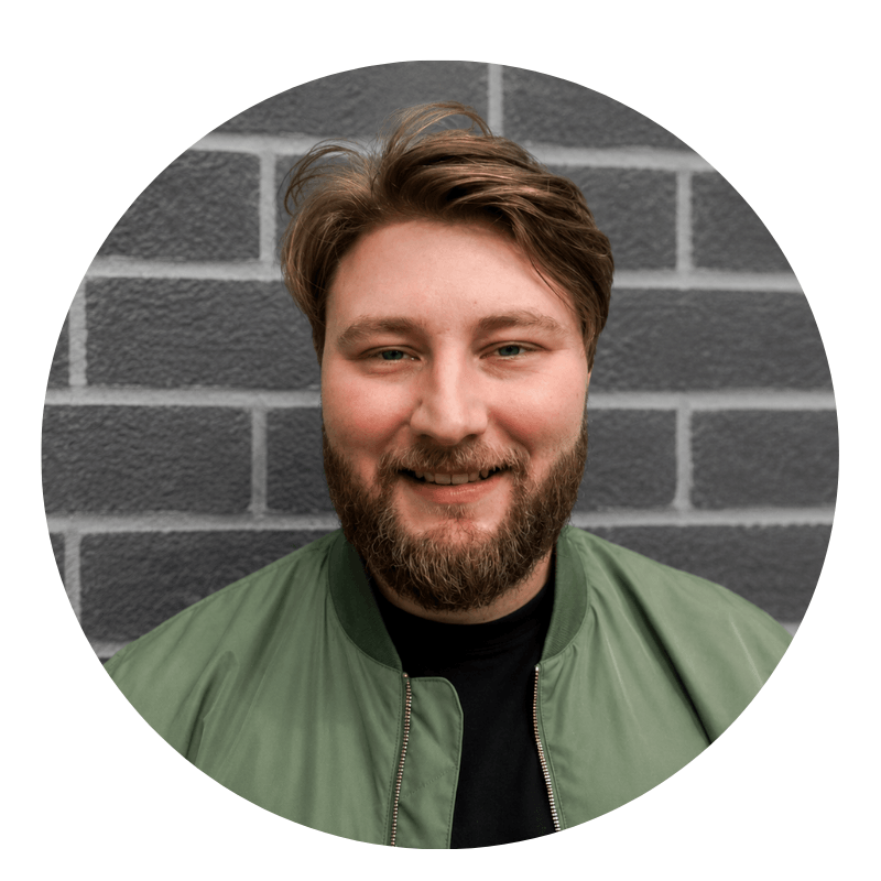 trevor-grimes-2026 (1) A man with light brown hair and a beard, wearing a green jacket over a black shirt, smiling in front of a gray brick wall.