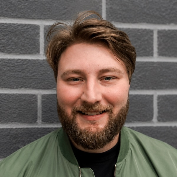 trevor-grimes A man with light brown hair and a beard smiles at the camera, standing in front of a gray brick wall and wearing a green jacket over a black shirt.