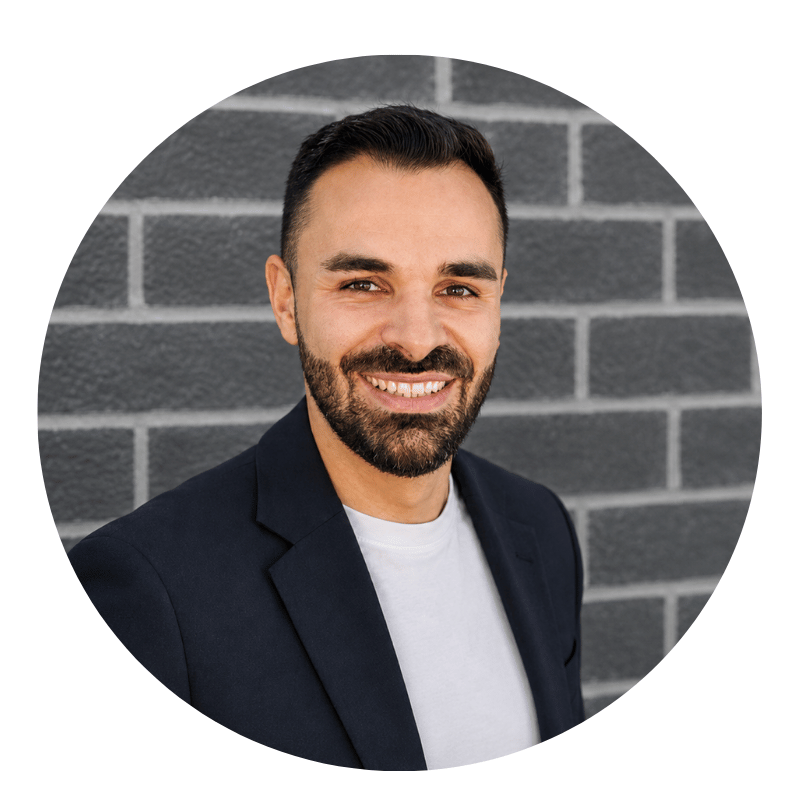 vlad-dordevic-2026 Smiling man with dark hair and beard, wearing a dark blazer and white shirt, standing in front of a gray brick wall.